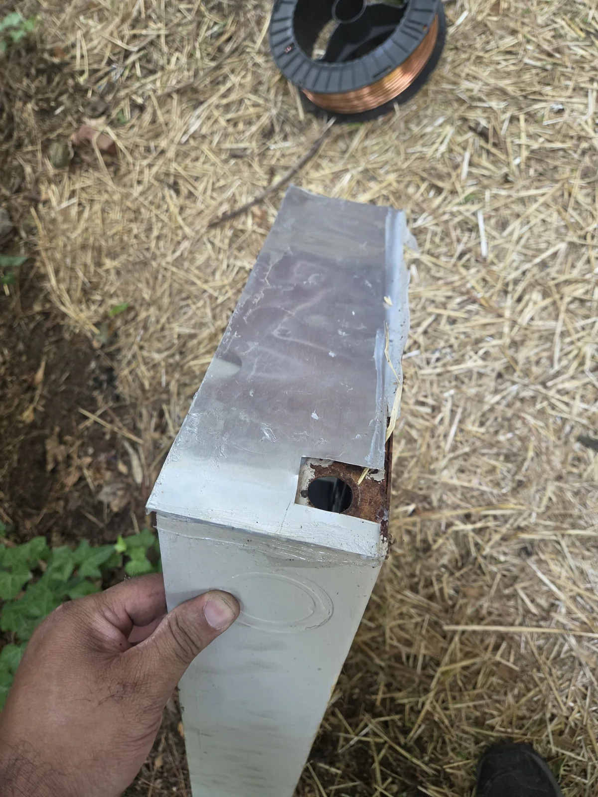 A hand holding an old electrical panel with a hole rusted through its side, outdoors on straw, in Oakwood, Raleigh